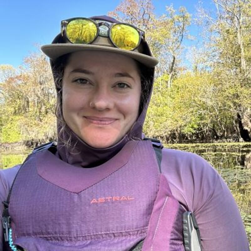 The image shows a woman wearing a purple life vest and a hat with sunglasses on top. She is outdoors, possibly kayaking or engaging in a water sport, with trees and water visible in the background. She is smiling and looking at the camera. The life vest has the word "ASTRAL" written on it.